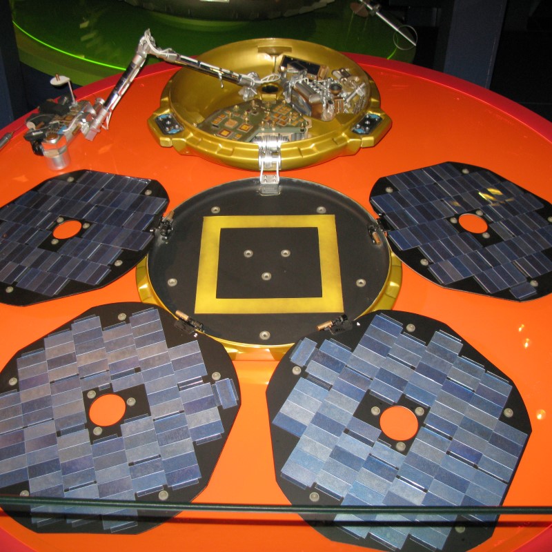 Photograph of Beagle 2 Mars Lander replica in the London Science Museum. The lander is composed 5 discs around a central disc. The discs around the outside have patches representing solar panels. A mechanical arm extends from the center.