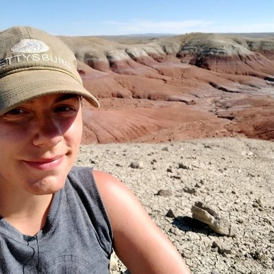 Alex Shupinski photographed at the Bighorn Basin of Wyoming. Alex wears a hat in the foreground, and the background is the red and sandy coloured basin with a blue sky.