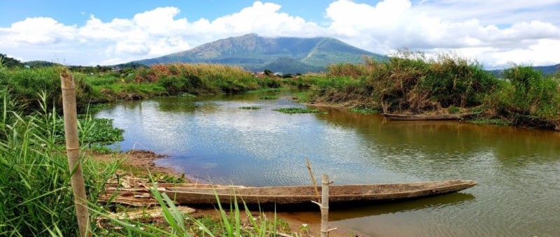 Photograph of Lake Buhi in Camarines Sur, Philippines, by Justin M. Bernstein. The photograph looks upriver along green banks to a mountain in the distance. In the foreground is a simple wooden boat, half on the water and half on the river bank.