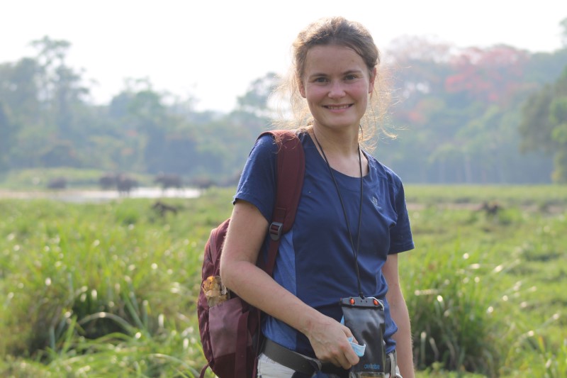 Photograph of Lara Nellissen standing in a grass field with a forest in the back ground, smiling at the camera. Taken by Paul Mboko.