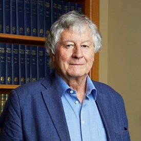 A white man with grey hair wearing a blue shirt and jacket, standing in front of a bookcase.