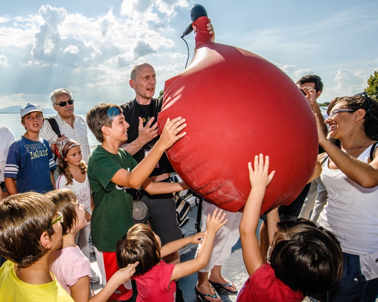 Man outdoors inflating a giant balloon using a hair dryer, with an audience of children