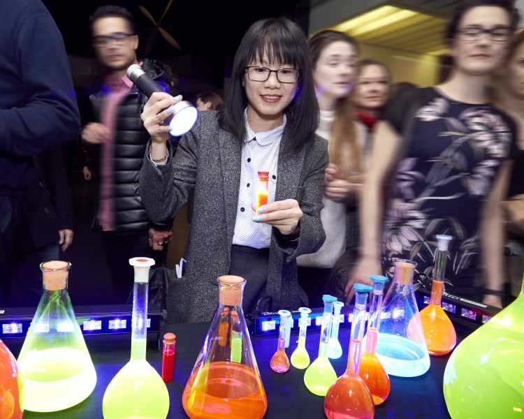 Woman shining a torch on a selection of conical flasks and test tubes full of coloured liquid that glows under the light